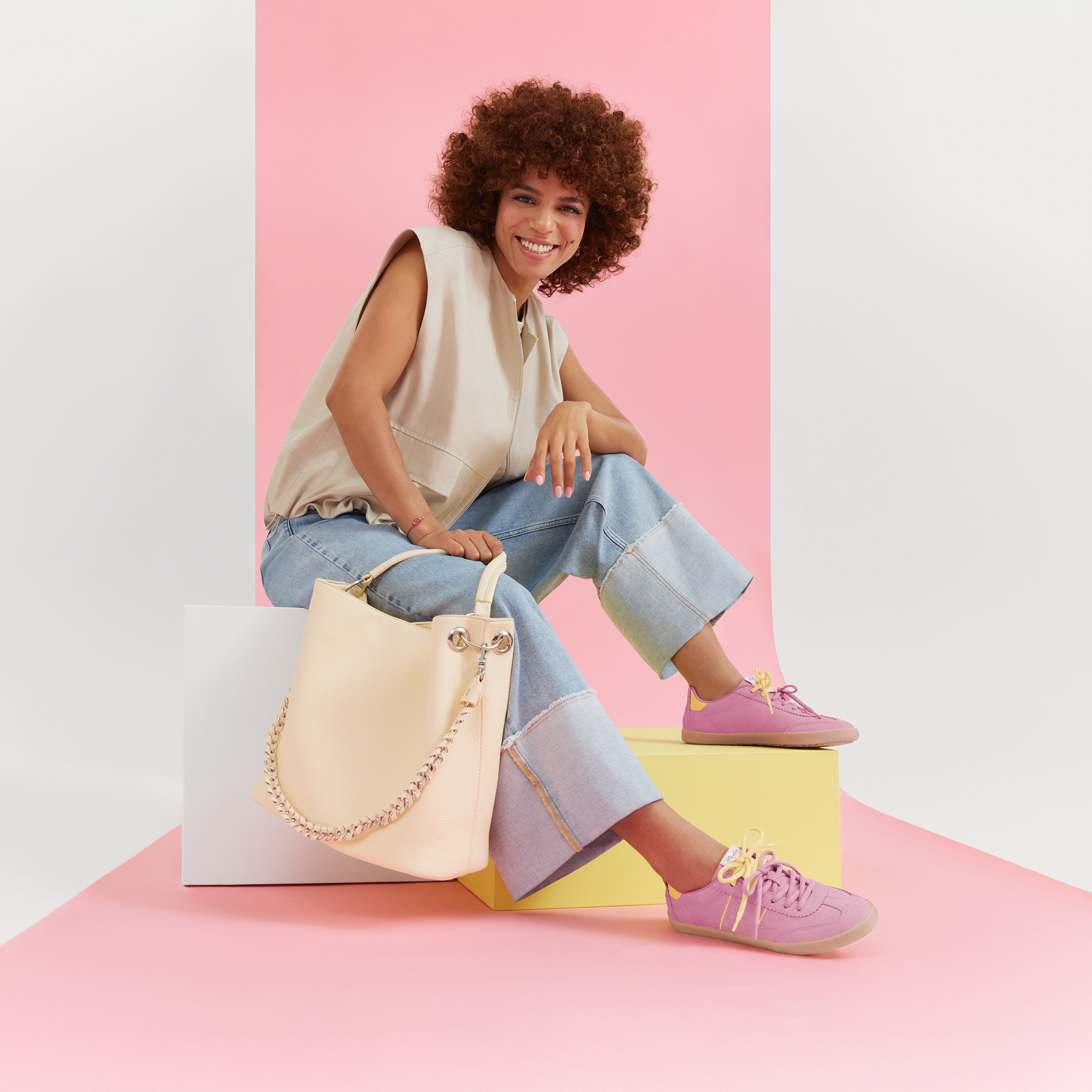 Outfit alla moda con top beige, jeans, sneakers rosa e borsa a mano color crema su sfondo pastello.