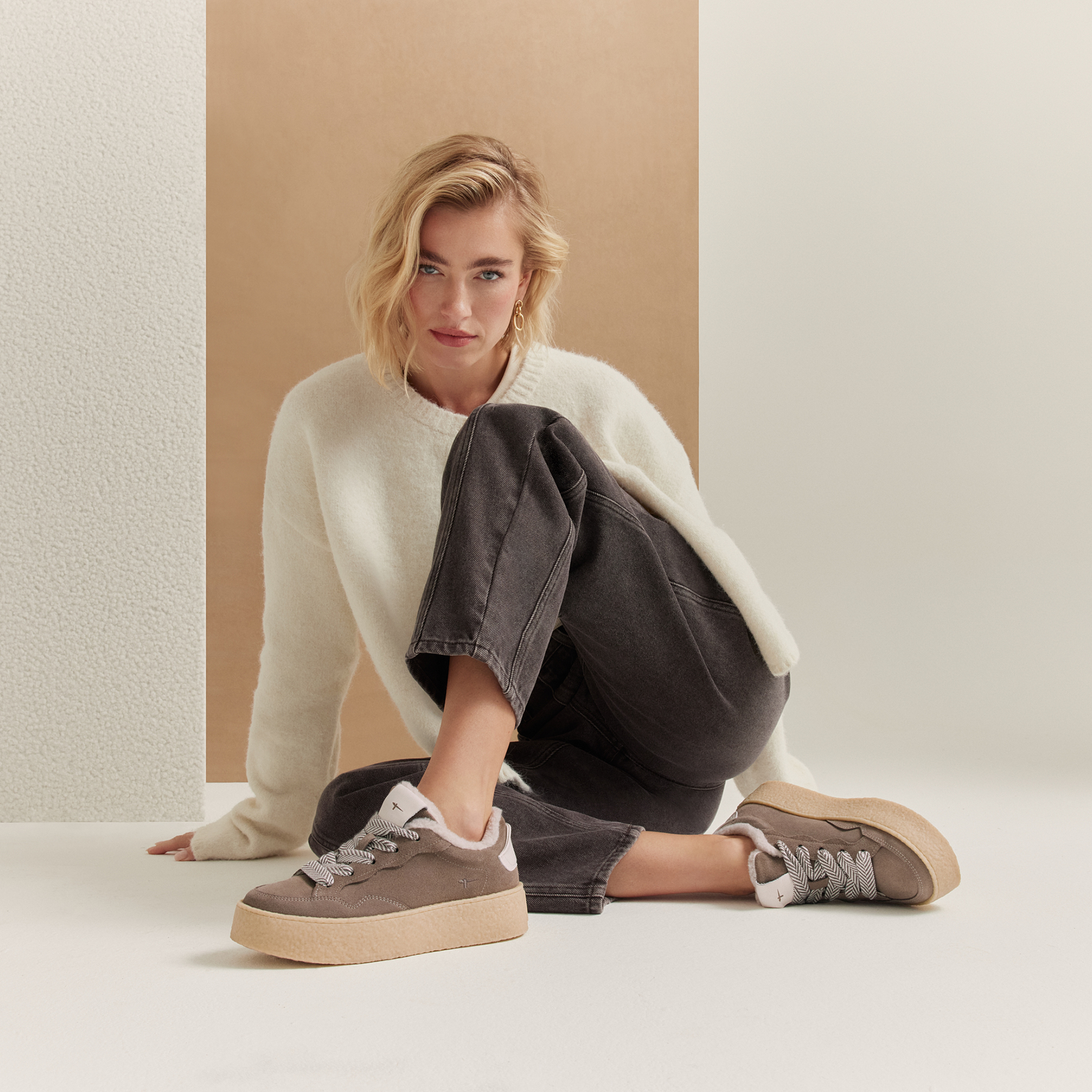 Frau mit blonden Haaren sitzt auf dem Boden, tr&auml;gt cremefarbenen Pullover, graue Jeans und braune Plateau-Sneaker.