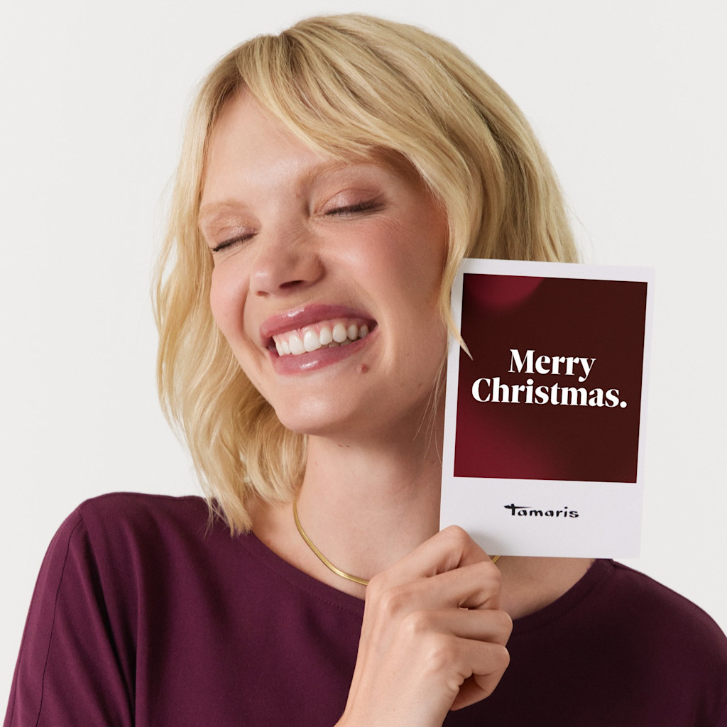 Lächelnde Person in bordeauxrotem Oberteil hält eine weiße Karte mit der Aufschrift "Merry Christmas" von Tamaris