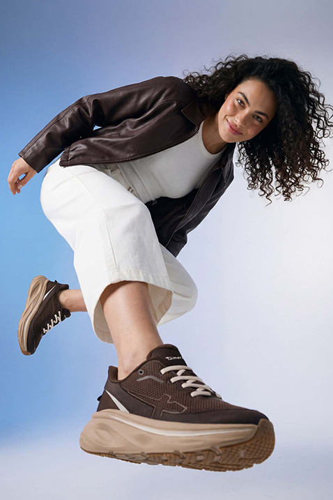 Femme aux cheveux boucl&eacute;s sautant en veste en cuir marron, pantalon blanc et sneakers Tamaris marron devant un fond bleu.