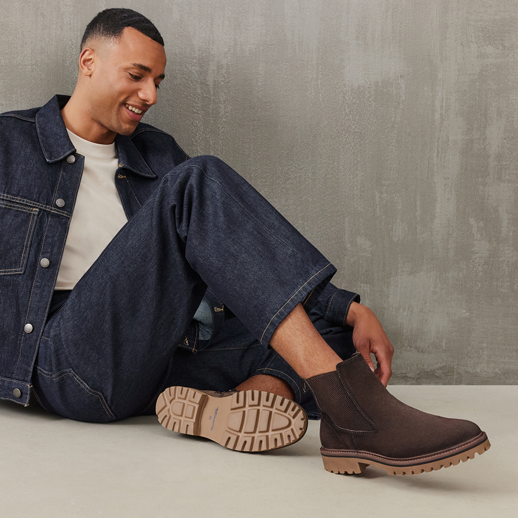 Man in een spijkerjas en jeans zit glimlachend op de grond en draagt bruine Chelsea‑boots met een lichte profielzool.