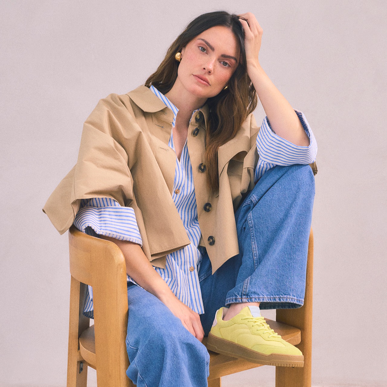 Modieuze outfit met een beige trenchcoat, gestreepte blouse, spijkerbroek en gele sportschoenen tegen een lichte achtergrond.