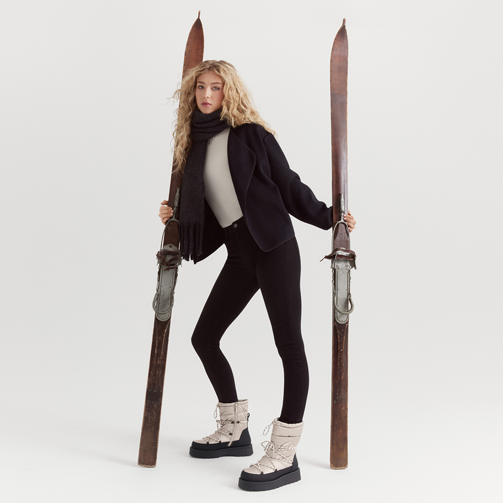 Modèle en tenue d'hiver noire avec des bottes blanches posant de manière dynamique entre des skis vintage sur fond blanc.
