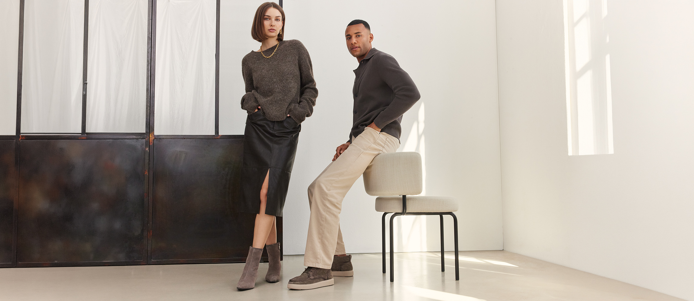 Woman in a grey jumper and black leather skirt with ankle boots, man in a dark shirt and beige trousers with trainers, sitting on a chair.