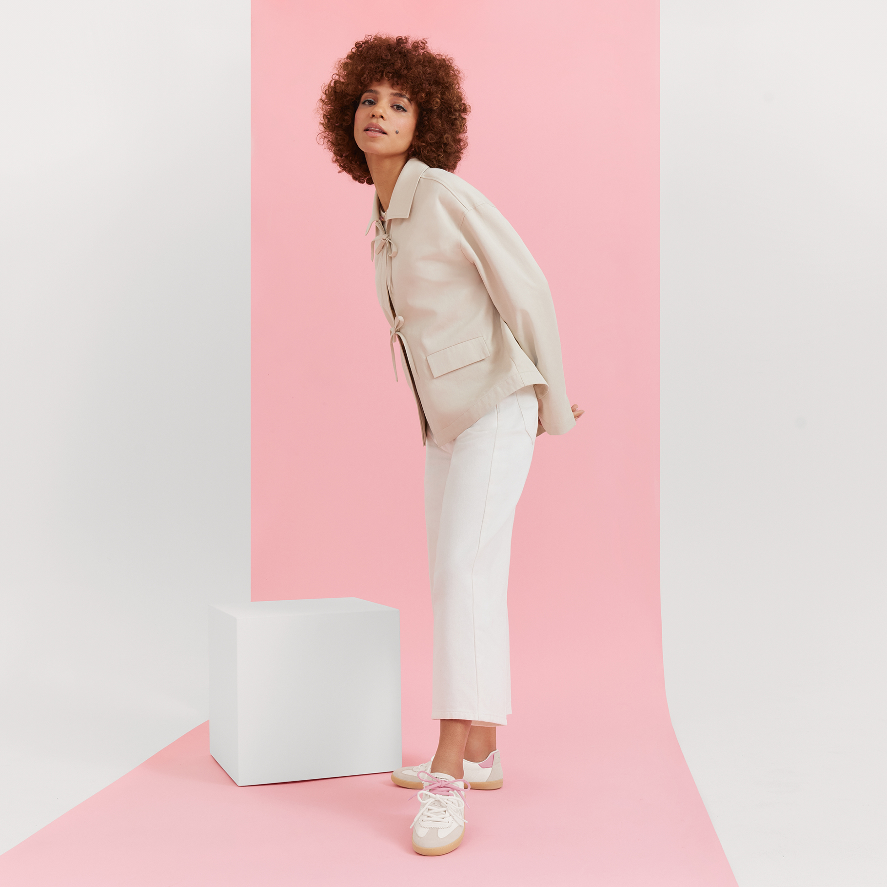 Modell in cremefarbenem Blazer und wei&szlig;er Hose vor rosa Hintergrund, mit lockiger Frisur und wei&szlig;en Sneakern.