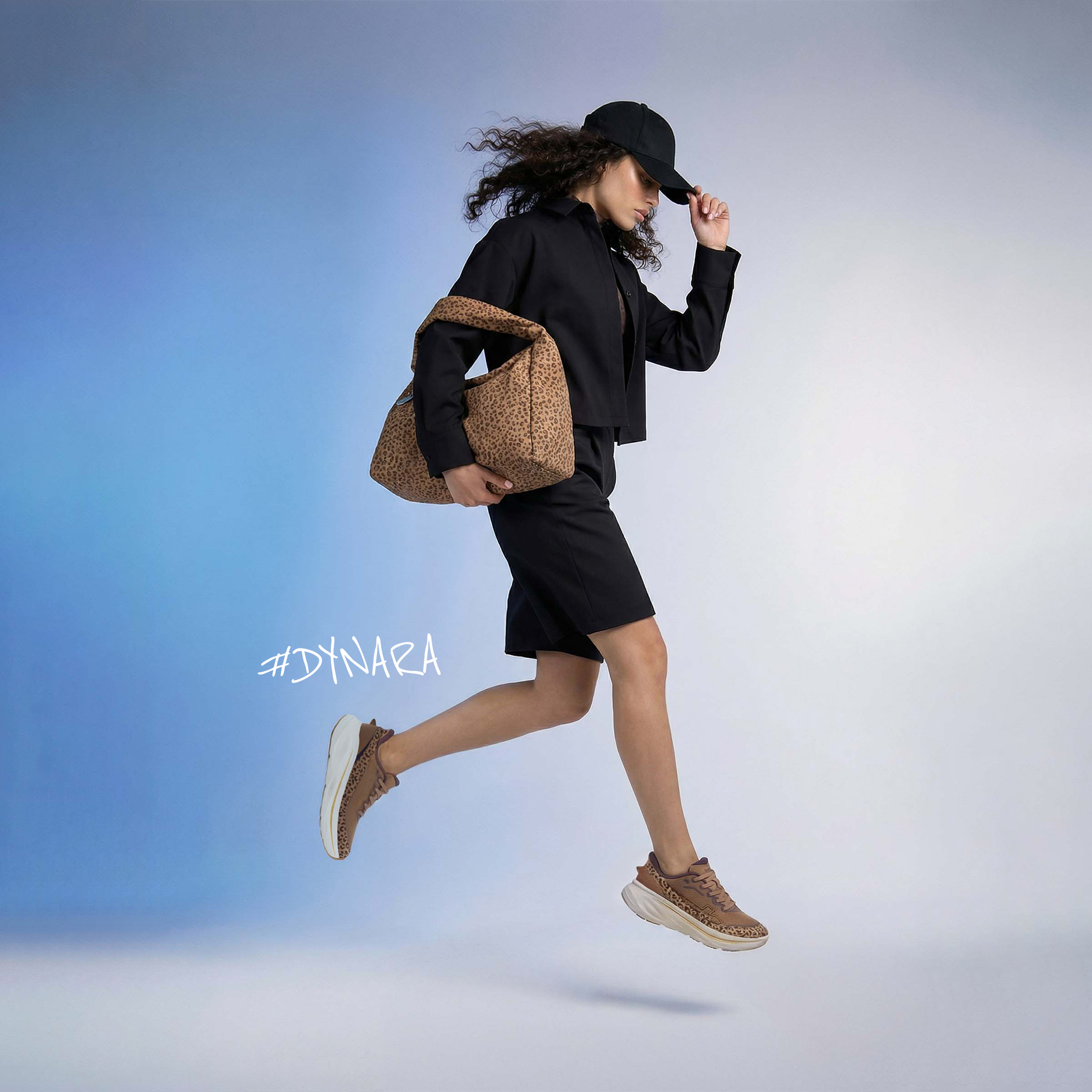 Donna con un outfit nero salta, indossa una borsa leopardata, scarpe da ginnastica e un berretto nero. Hashtag #DYNARA nell'immagine.