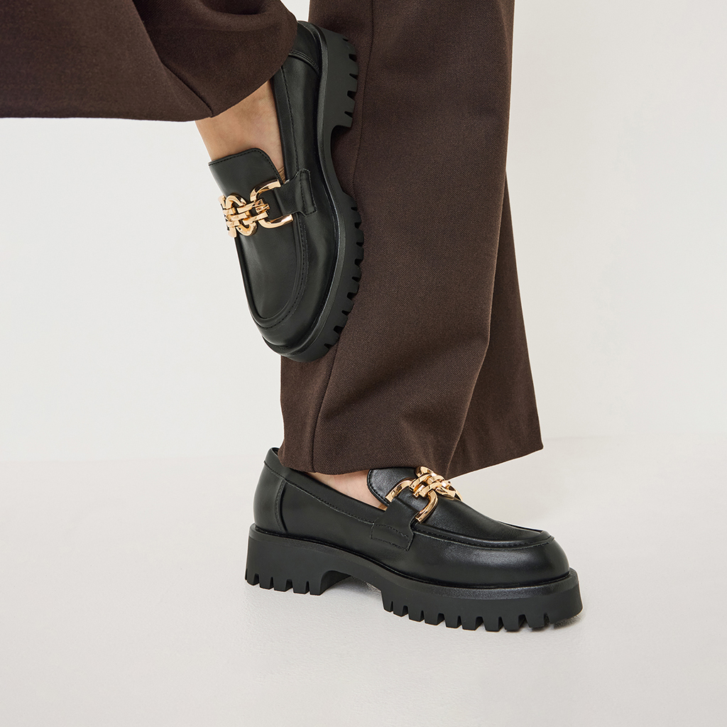 Schwarze Plateau-Loafer mit goldener Kettenverzierung, getragen zu einer braunen Hose vor wei&szlig;em Hintergrund.