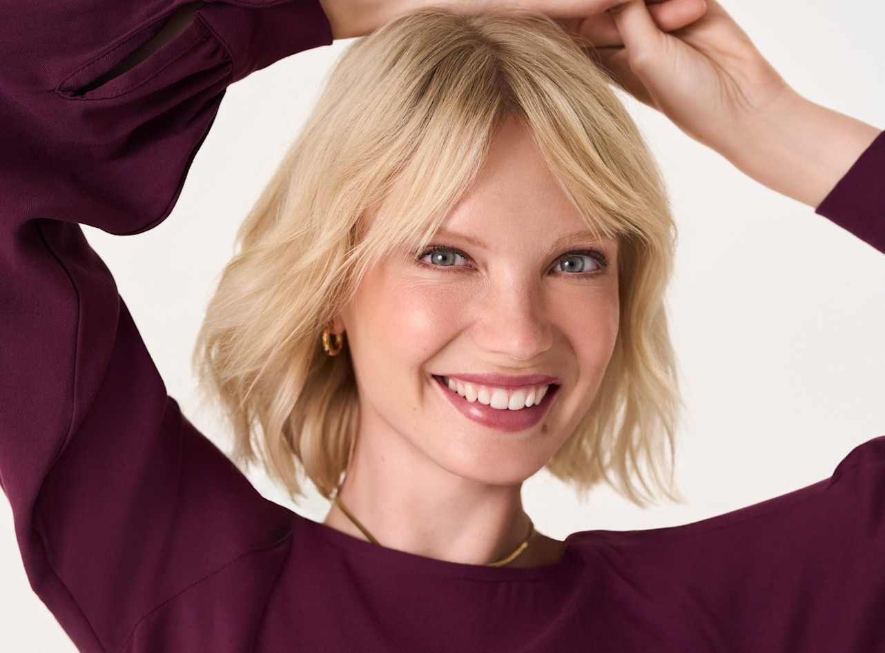 Nahaufnahme einer l&auml;chelnden Person mit blonden Haaren in einem burgunderroten Pullover vor wei&szlig;em Hintergrund