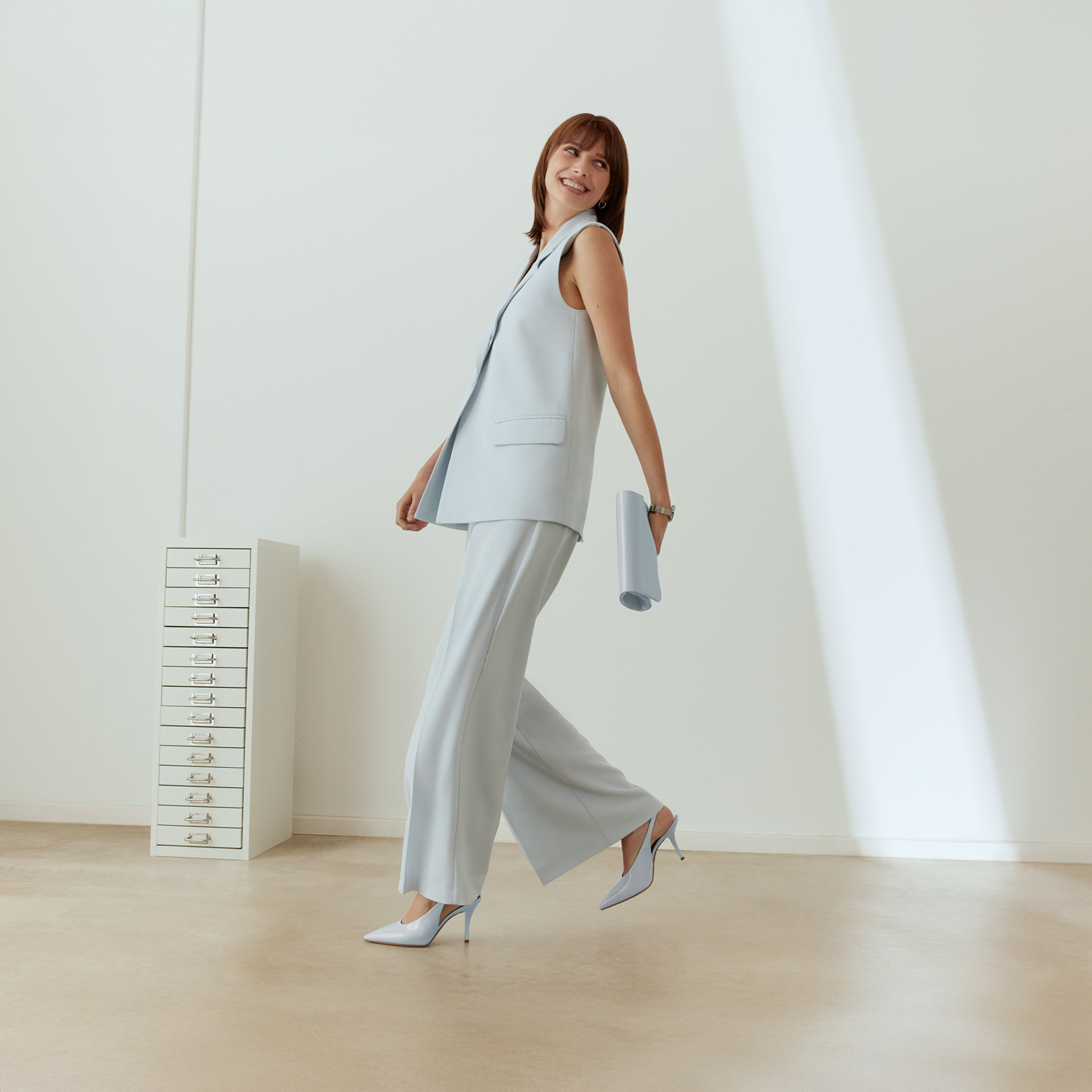 Donna in abito business azzurro chiaro con gilet senza maniche e pantaloni ampi, sorridente in posa dinamica davanti a una parete bianca.