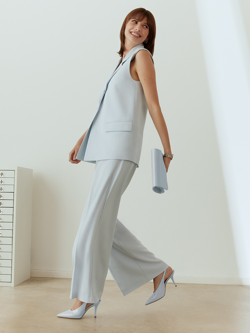 Donna in abito business azzurro chiaro con gilet senza maniche e pantaloni ampi, sorridente in posa dinamica davanti a una parete bianca.