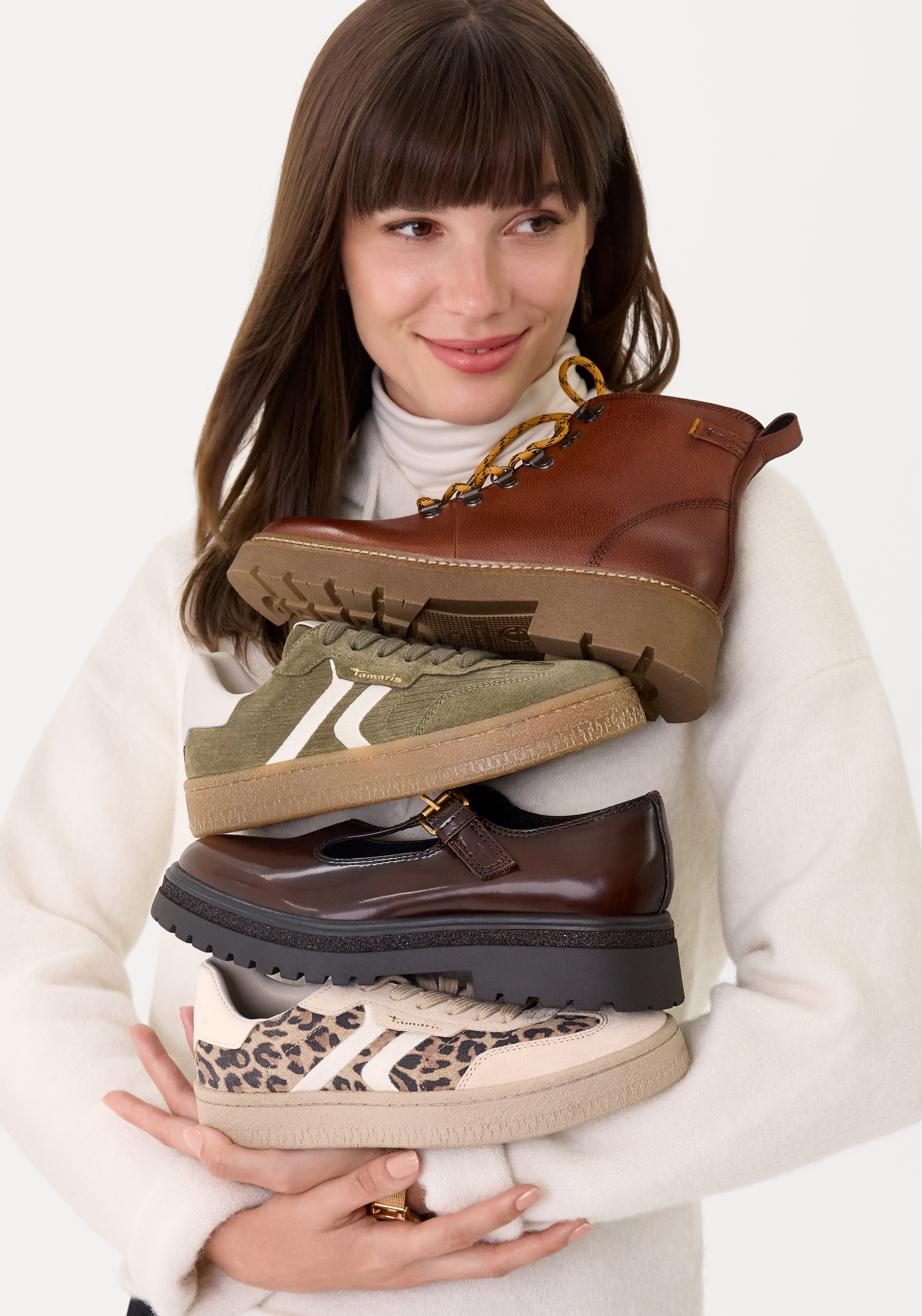 Eine Frau in weißem Pullover hält verschiedene Schuhe, darunter Stiefel, Sneaker und Loafer in Braun- und Leopardentönen.
