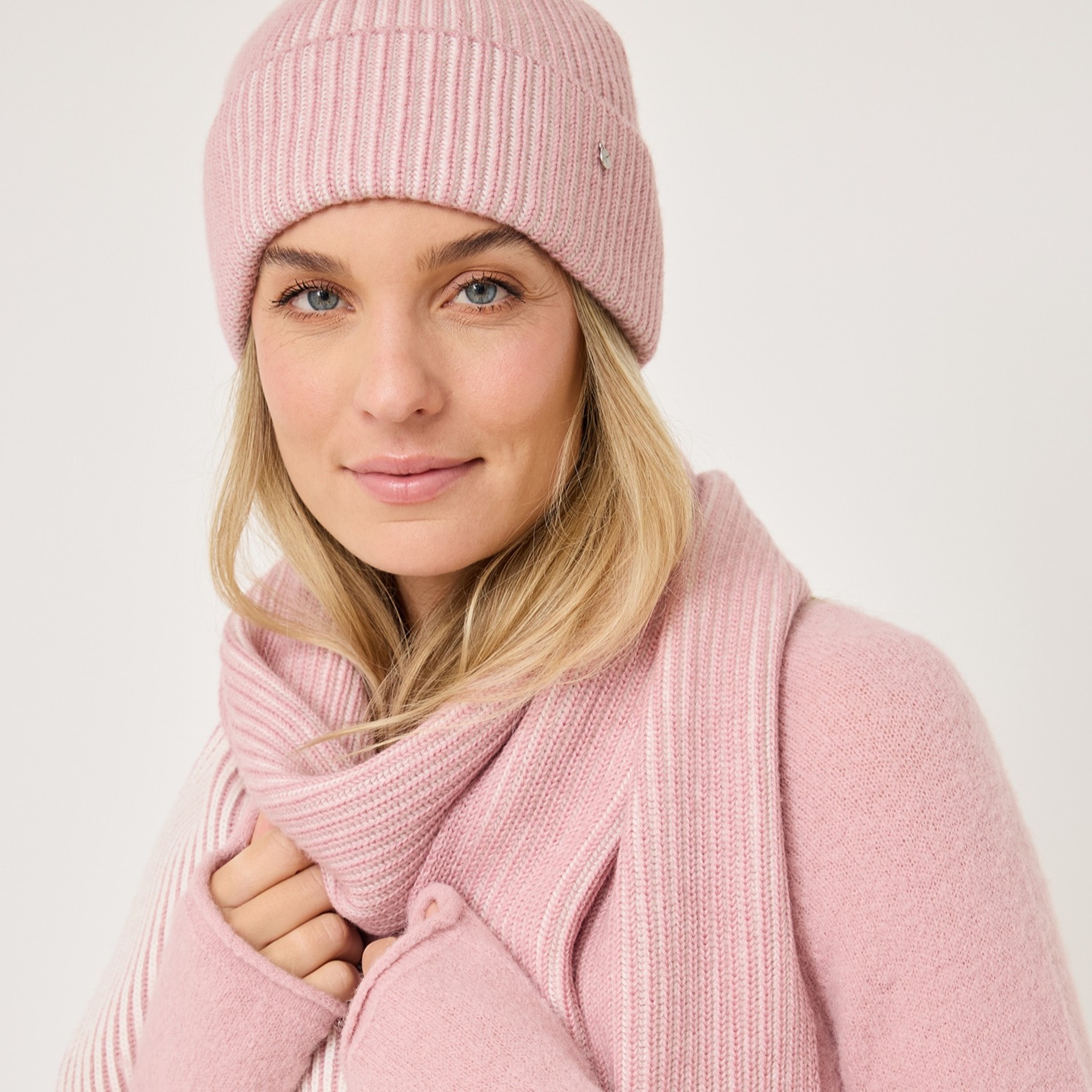 Close-up of a person in a pink knitted hat and sweater against a white background, with blond hair and natural make-up.