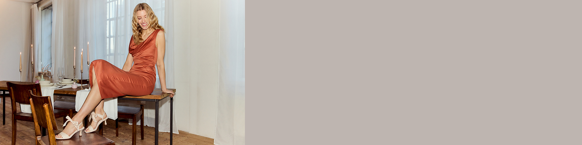 Donna in abito di seta color ruggine seduta con eleganza su un tavolo di legno, indossa sandali beige con fiocco.