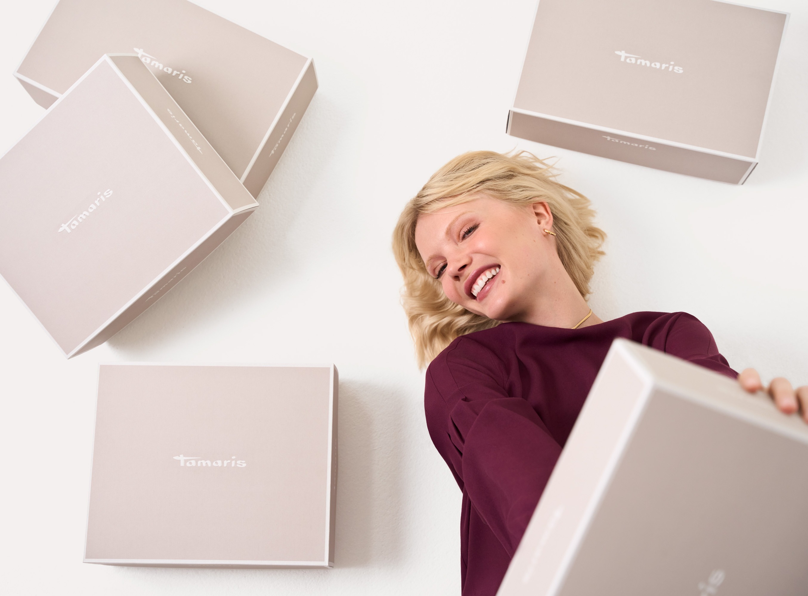 L&auml;chelnde Frau in bordeauxrotem Pullover zwischen gestapelten wei&szlig;en Tamaris-Schuhkartons vor hellem Hintergrund