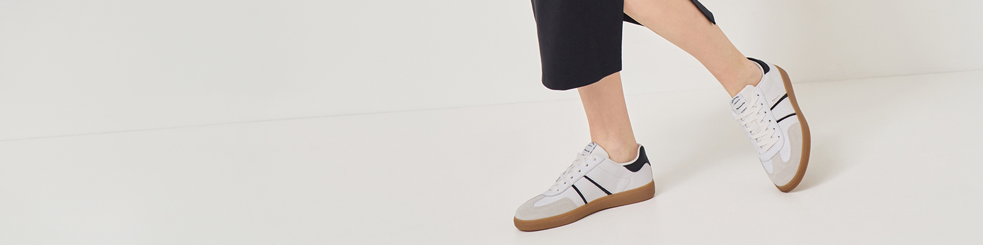 White sneakers with a brown rubber sole, worn with a black skirt, against a light background.