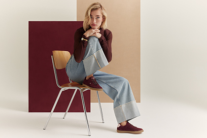Kvinna sitter p&aring; en tr&auml;stol, b&auml;r bordeauxr&ouml;d tr&ouml;ja, vida jeans och r&ouml;da trainers mot beige‑r&ouml;d bakgrund.