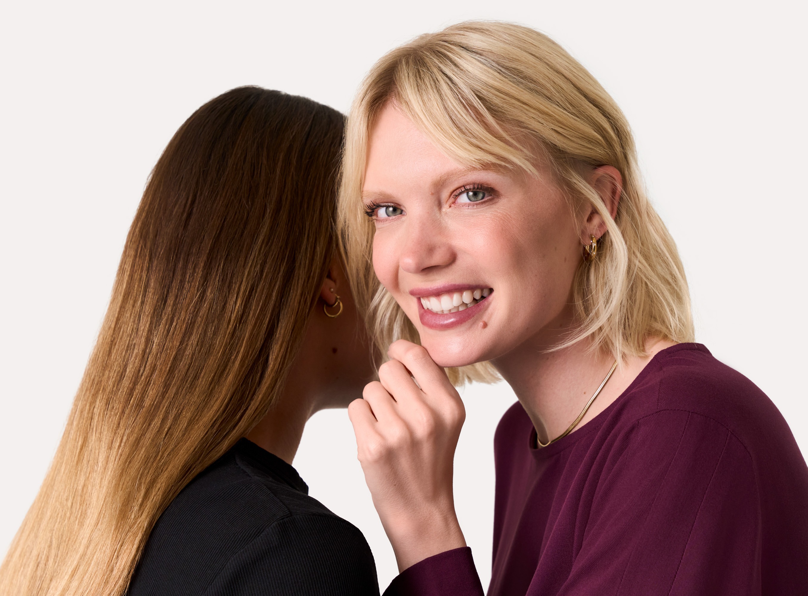 Eine junge Frau mit blonden Haaren in einem lila Oberteil l&auml;chelt vor wei&szlig;em Hintergrund in die Kamera.
