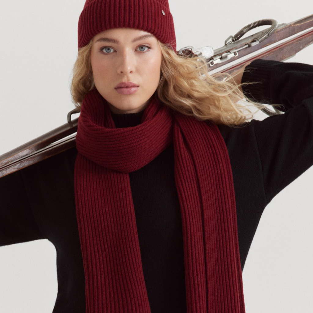 Frau mit schwarzem Strickkleid, weinrotem Schal und M&uuml;tze tr&auml;gt Ski &uuml;ber der Schulter vor wei&szlig;em Hintergrund