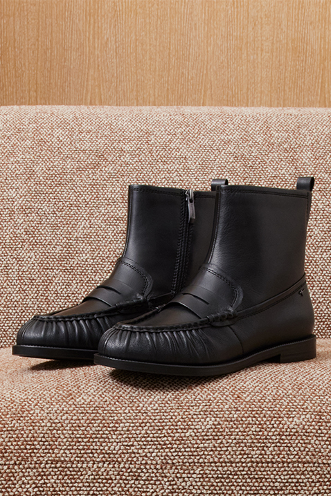 Schwarze Leder-Stiefeletten mit Falten-Details entlang der Schuhspitze, auf einem beigen Stoffsofa platziert.