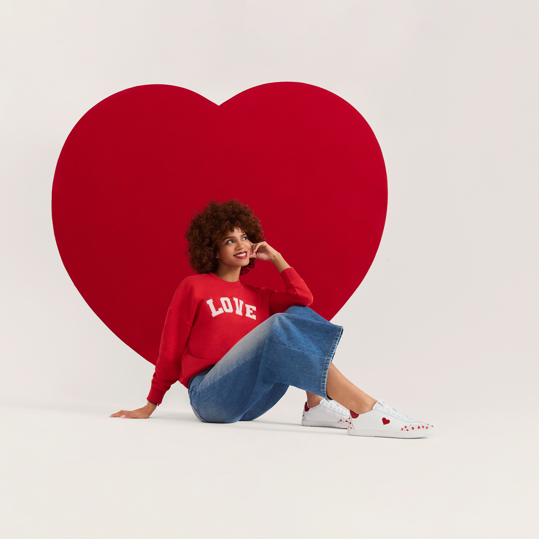 Persona sorridente con maglione rosso LOVE e jeans davanti a uno sfondo di cuore rosso, con sneaker bianche e capelli ricci.