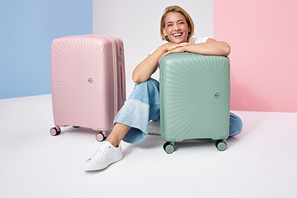 Donna sorridente seduta tra due valigie, una rosa e una verde menta, davanti a uno sfondo color pastello.