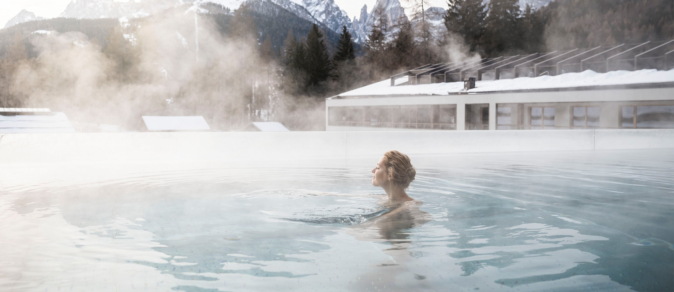 Frau entspannt im dampfenden Au&szlig;enpool eines Wellnesshotels, umgeben von schneebedeckten Bergen im Winter.