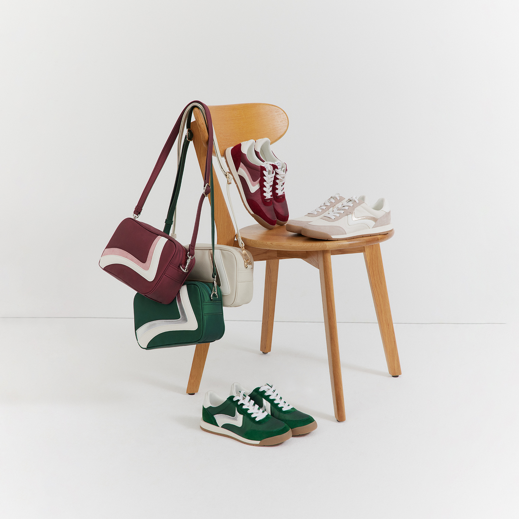 Sporty sneakers in green, red and beige with matching handbags, arranged around a wooden chair on a white background.