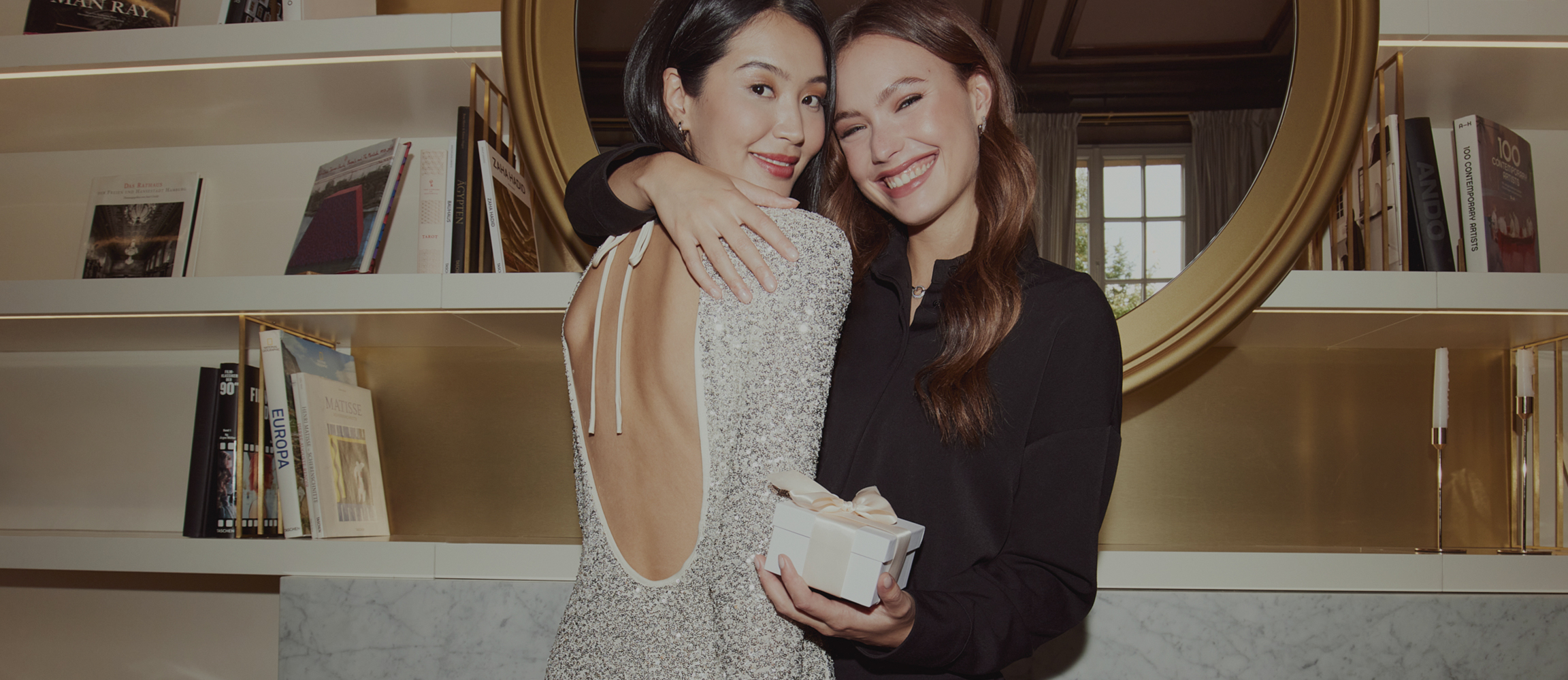 Two women are smiling at the camera in front of an elegant mirror with a golden frame, one is wearing a white dress, the other a black one.