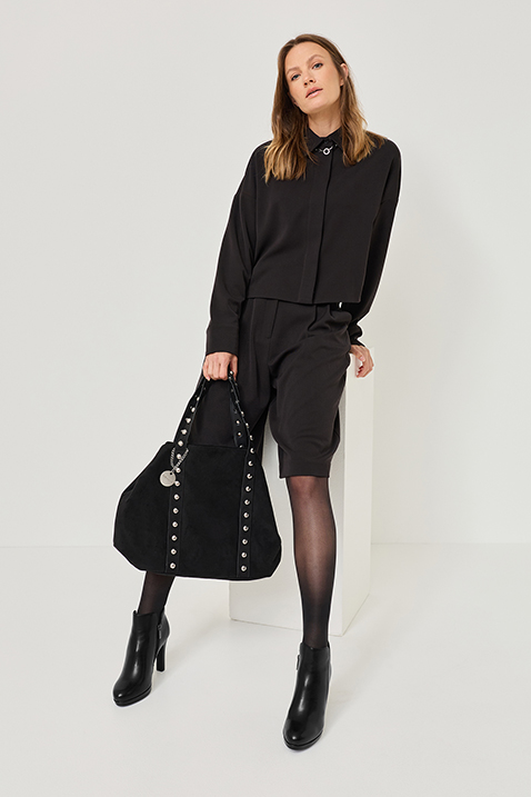 Vrouw in elegante zwarte zakelijke outfit met blouse, korte broek en laarzen en zwarte tas poseert voor een witte achtergrond