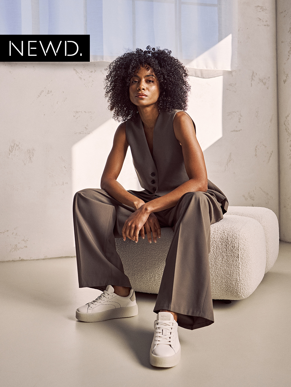 Modèle en tenue d'affaires grise avec des baskets blanches assis sur un banc blanc, lumière naturelle entrant par les fenêtres.