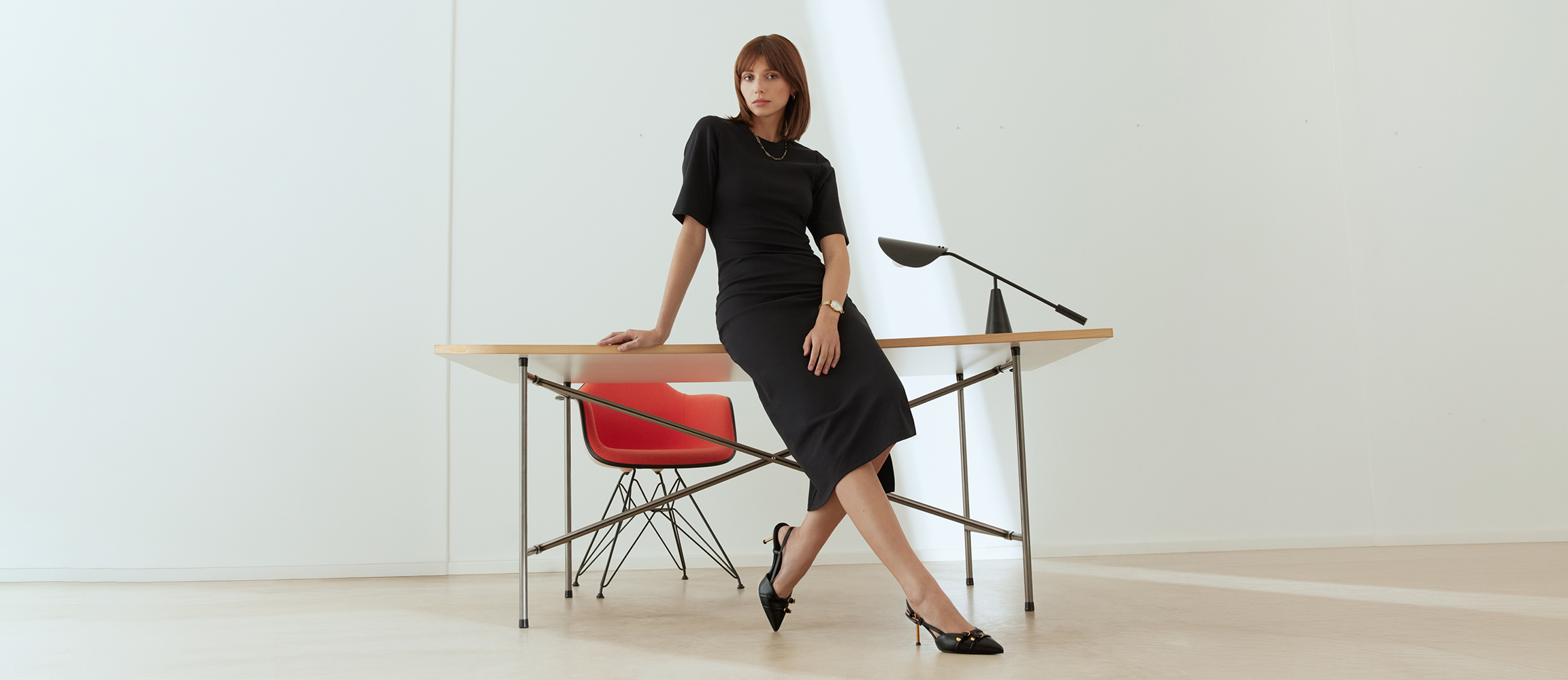 Eine Frau in schwarzem Kleid lehnt an einem modernen Schreibtisch mit rotem Stuhl in einem minimalistisch eingerichteten, hellen Raum.