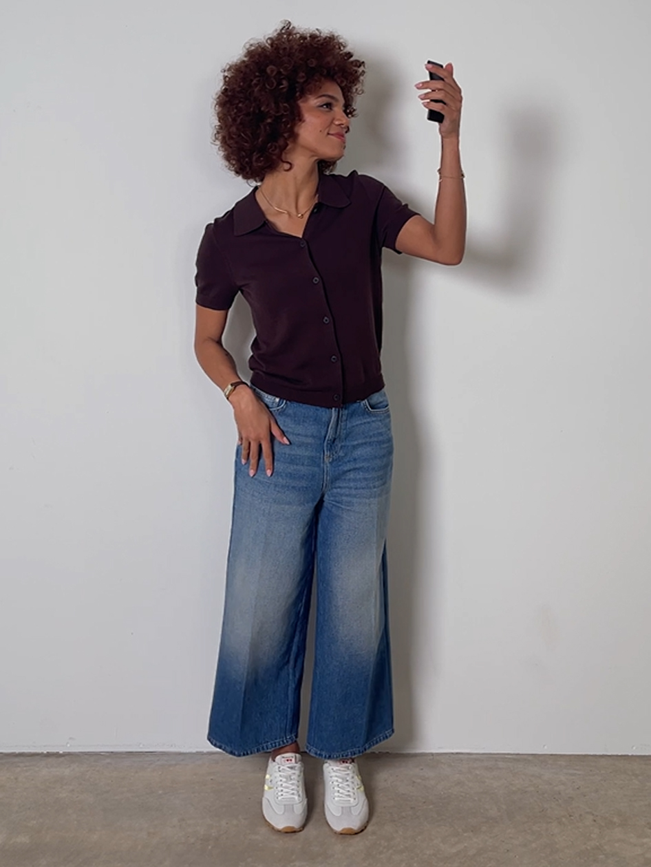 Femme aux cheveux boucl&eacute;s prenant un selfie, portant un polo marron, un jean bleu ample et des sneakers blanches.