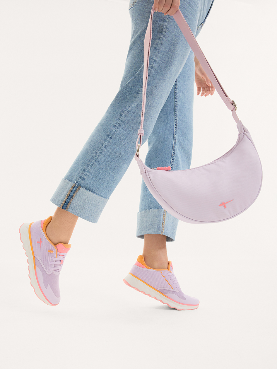 Person i jeans b&auml;r lila sneakers med orange detaljer och en halvm&aring;neformad crossbody‑v&auml;ska i lila.
