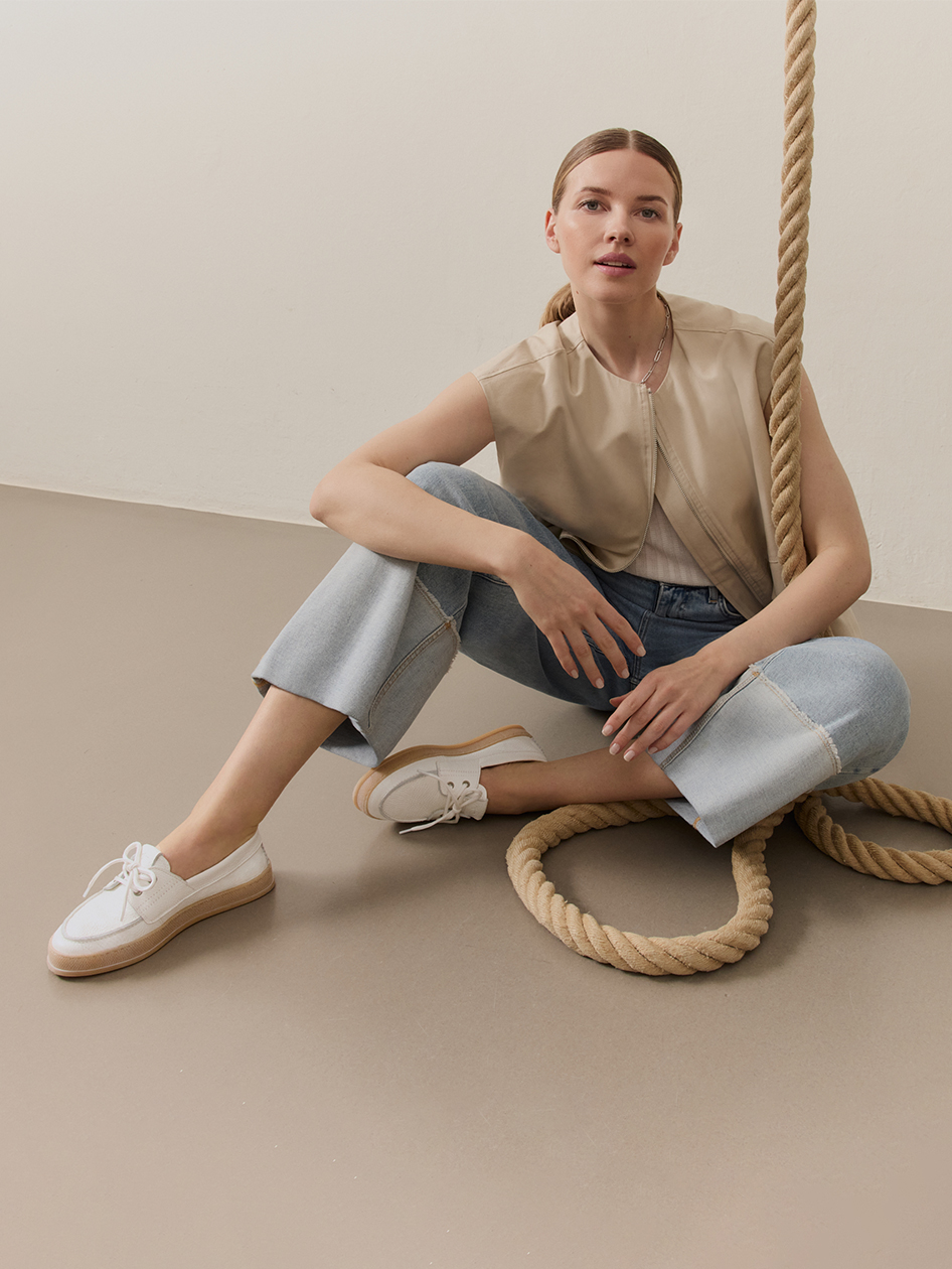 Donna con gilet beige e jeans seduta rilassata a terra, circondata da una grossa corda, indossa boat shoes bianche