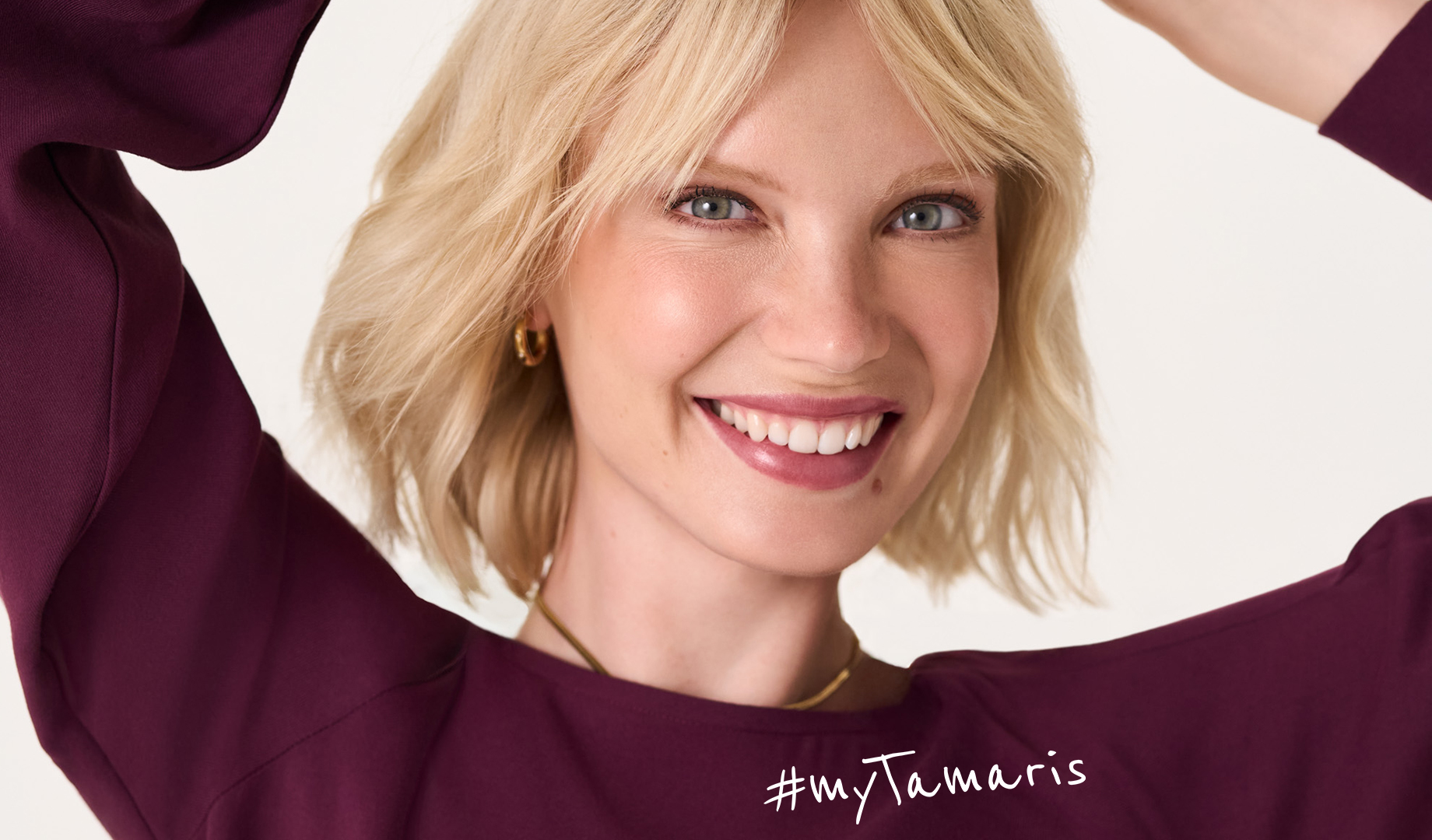 Nahaufnahme einer l&auml;chelnden Person mit blonden Haaren und blauen Augen in einem dunkelvioletten Oberteil vor wei&szlig;em Hintergrund.