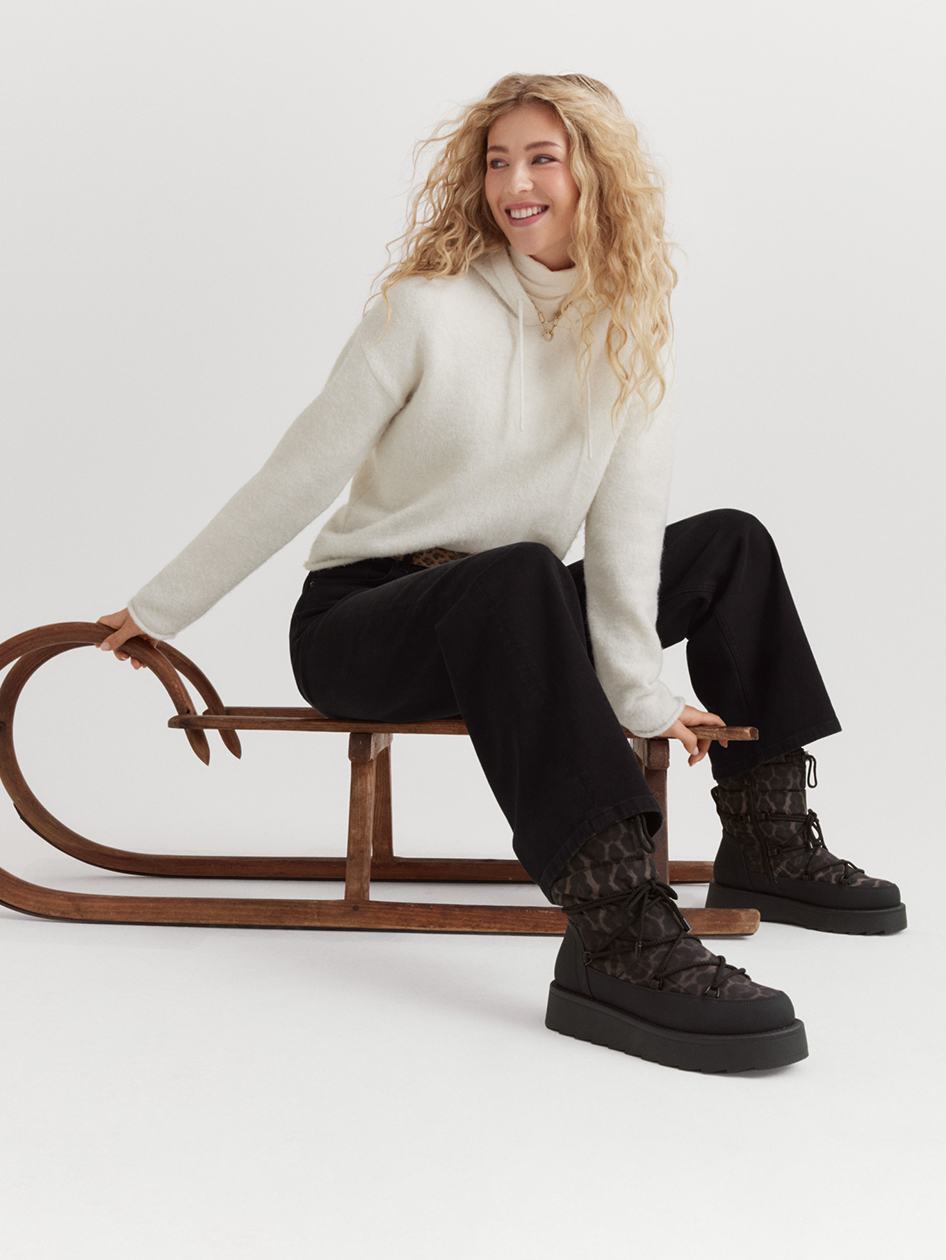 Vrouw in crèmekleurige trui en zwarte broek zit glimlachend op een houten slee, draagt zwarte winterlaarzen.