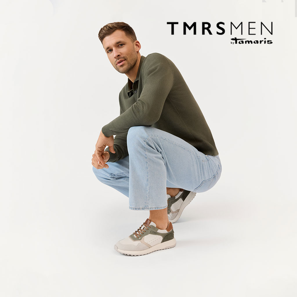 Man in olijfgroene trui en lichte jeans poseert dynamisch in hurkzit, draagt beige-groene sneakers op een witte achtergrond.
