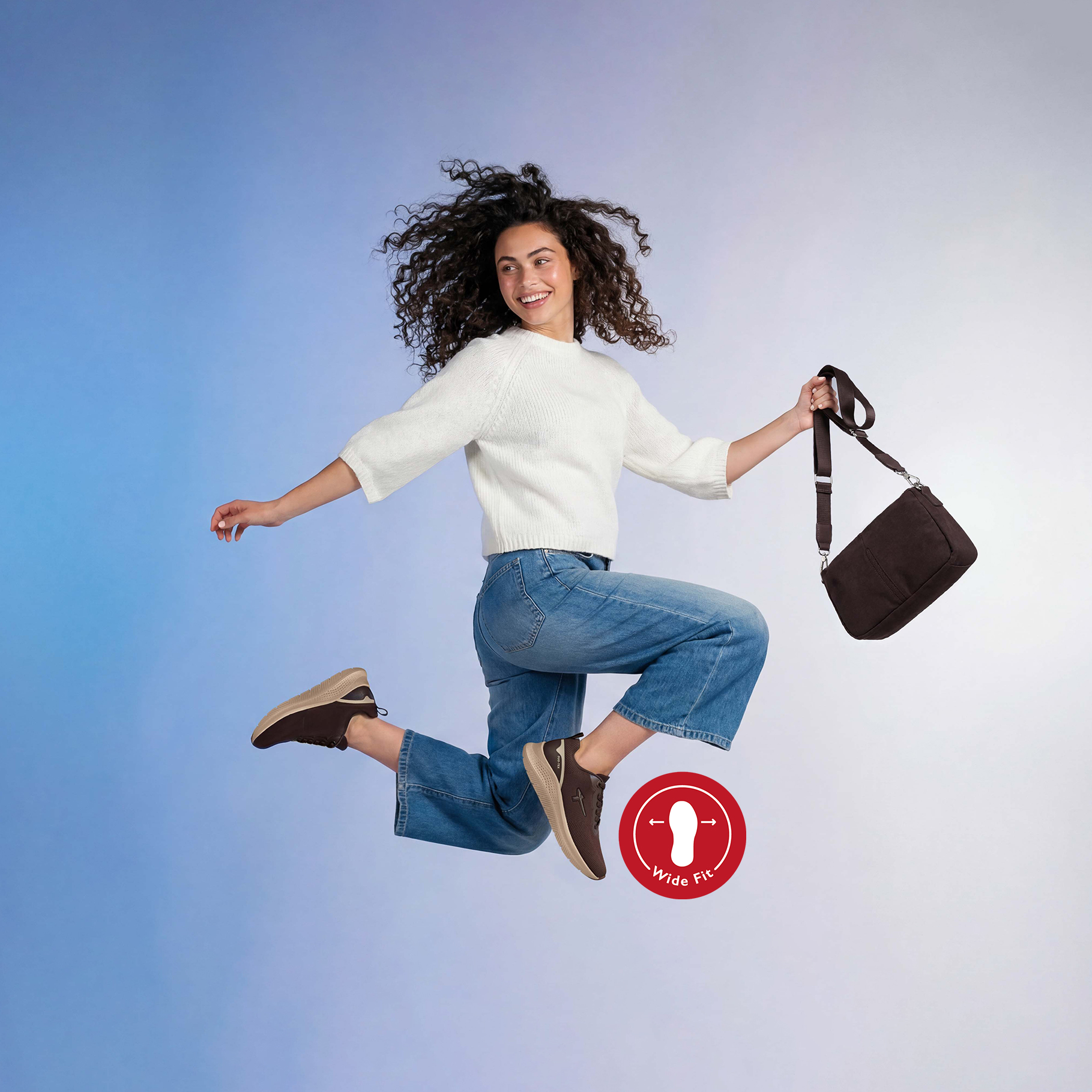 Donna salta allegramente in aria, indossa un maglione bianco, jeans, sneakers marroni (calzata ampia) e tiene in mano una borsa marrone.