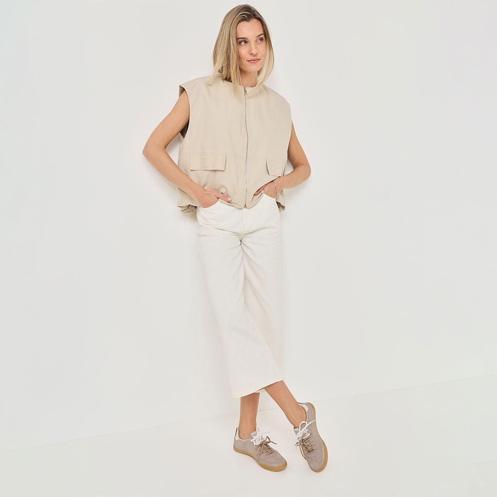 Model in beige mouwloze gilet, witte midirok en beige sneakers poseert voor een witte achtergrond.