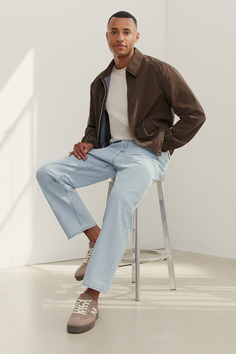 Mand sidder p&aring; en skammel, if&oslash;rt brun jakke, hvid T-shirt, lysebl&aring; jeans og beige sneakers foran lys baggrund.