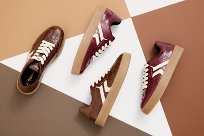 Quattro scarpe da ginnastica marroni con strisce bianche e suola in gomma, disposte in diagonale su uno sfondo bianco e marrone.