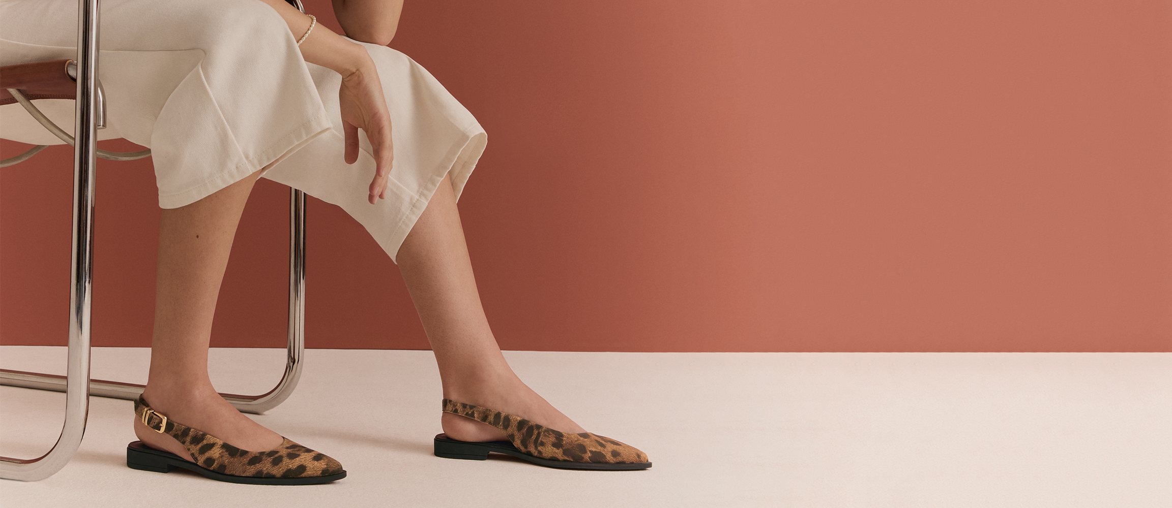 Vrouw zit op een stoel en draagt platte slingback‑schoenen met luipaardprint en een witte broek tegen een terracottakleurige achtergrond.