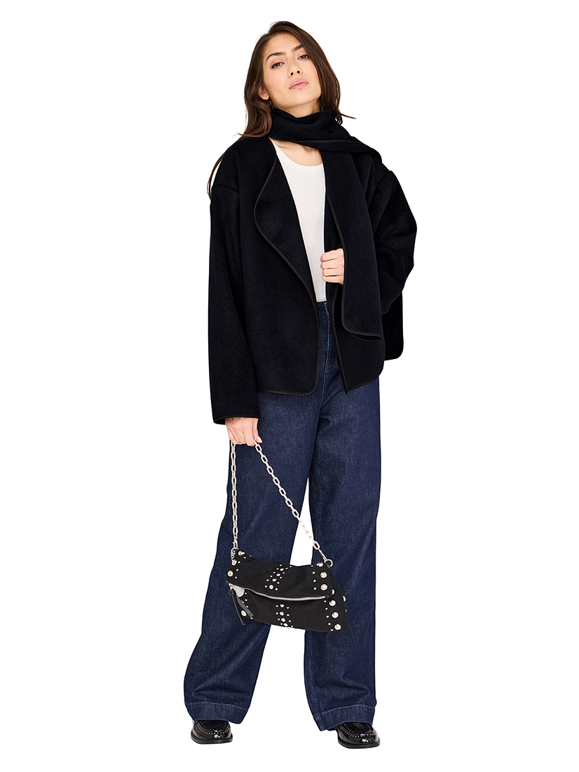 Modell in lässigem Outfit mit schwarzer Jacke, weiter Jeans und verzierter Handtasche mit Kettenriemen vor weißem Hintergrund.