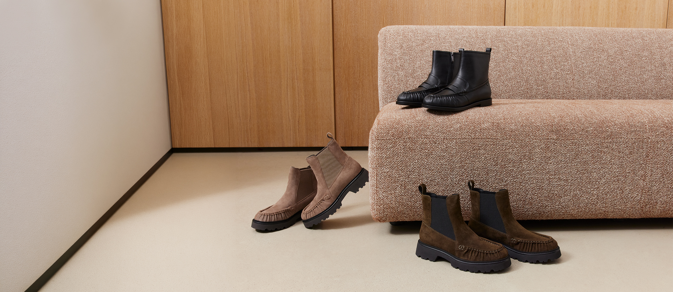 Drei Paar Chelsea-Boots in Schwarz, Taupe und Dunkelbraun, arrangiert auf dem Boden und einem beigen Sofa vor Holzwand.