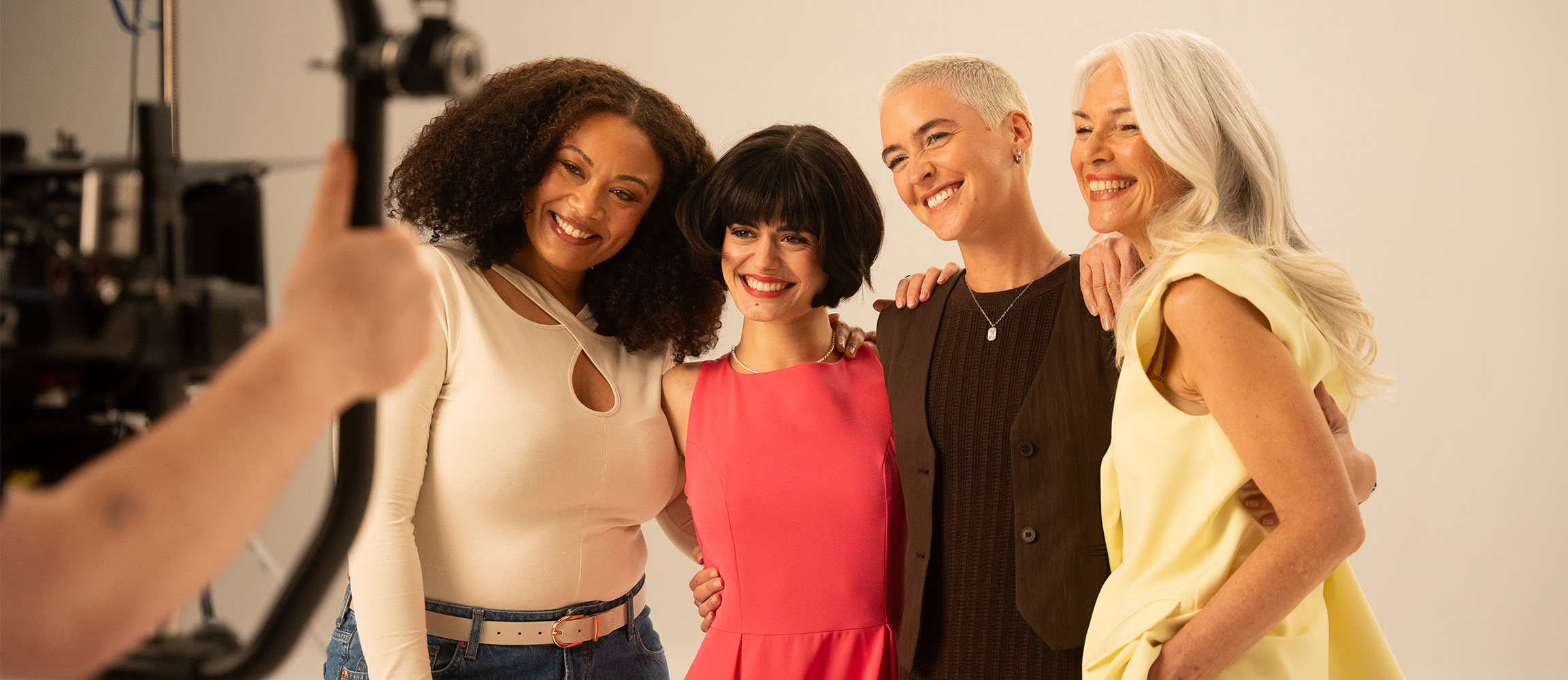 A diverse group of smiling women stands together in front of a light background during a studio photoshoot.