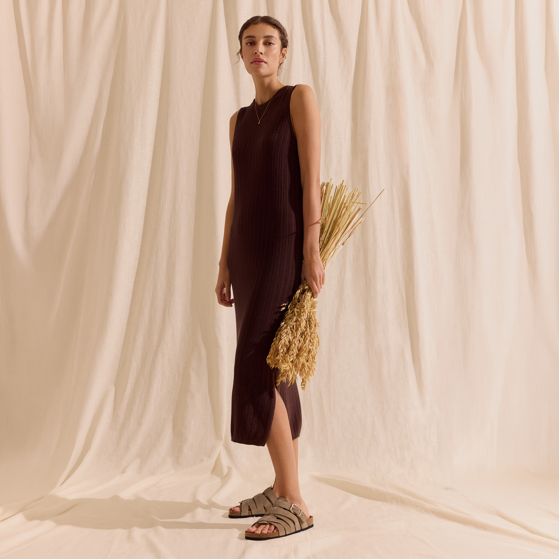 Femme en robe midi sans manches marron fonc&eacute; et sandales beiges devant un fond en tissu cr&egrave;me.