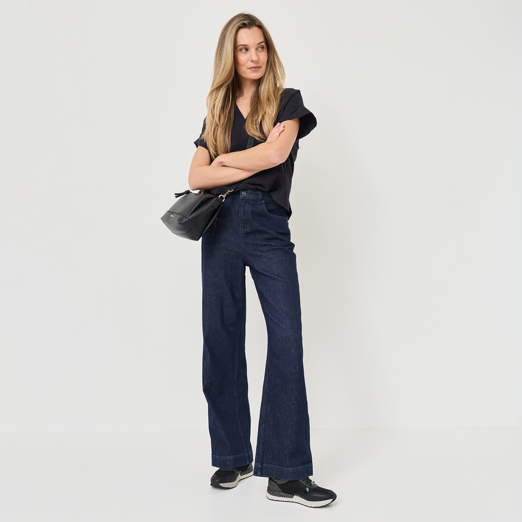 Kvinna med blont h&aring;r b&auml;r svart T-shirt, m&ouml;rkbl&aring; jeans och svarta sneakers och h&aring;ller en svart handv&auml;ska.