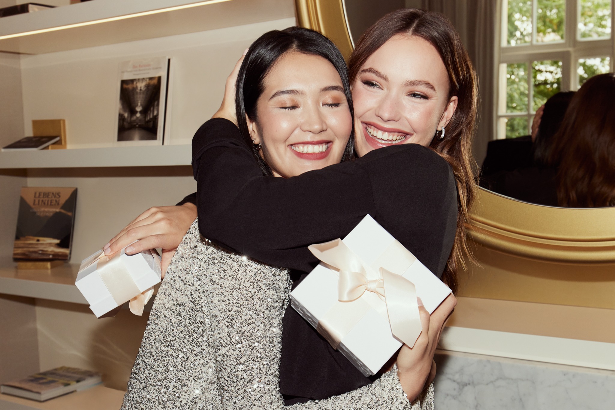 Due amiche si abbracciano con gioia, tengono in mano scatole regalo bianche con fiocchi, una indossa un abito scintillante, l'altra nero.