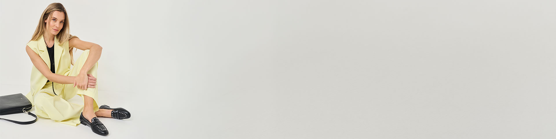 Žena ve světle žlut&eacute;m outfitu sed&iacute; na zemi a m&aacute; na sobě čern&eacute; loafers s cvoky. Vedle n&iacute; lež&iacute; čern&aacute; kabelka.