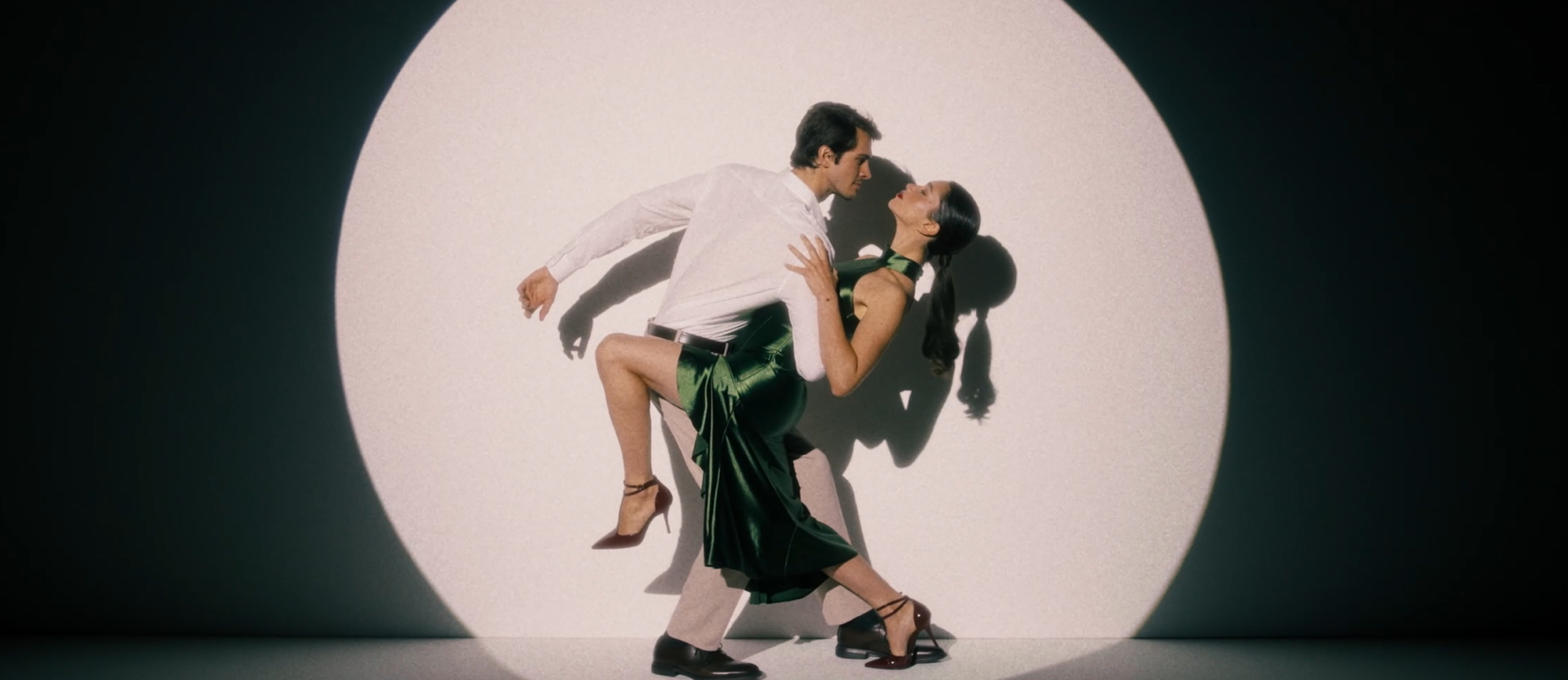 A couple dances elegantly in front of a round spotlight. The man is wearing white, the woman a green satin dress with high heels.
